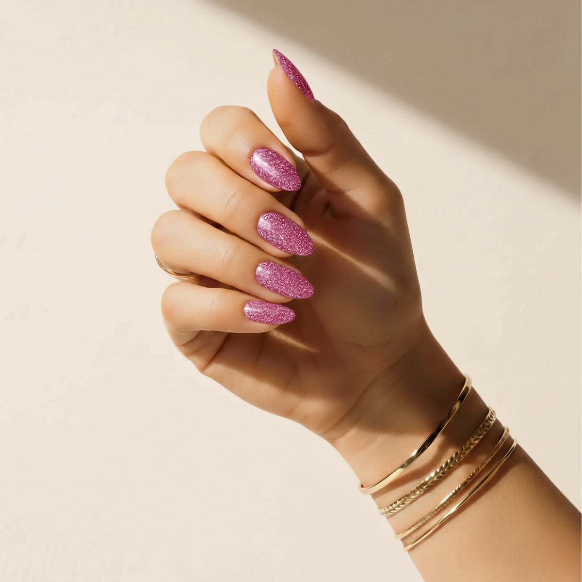 Main portant les autocollants pour ongles à paillettes roses Glitter Bug Delune et des joncs dorés devant un mur de plâtre chaud