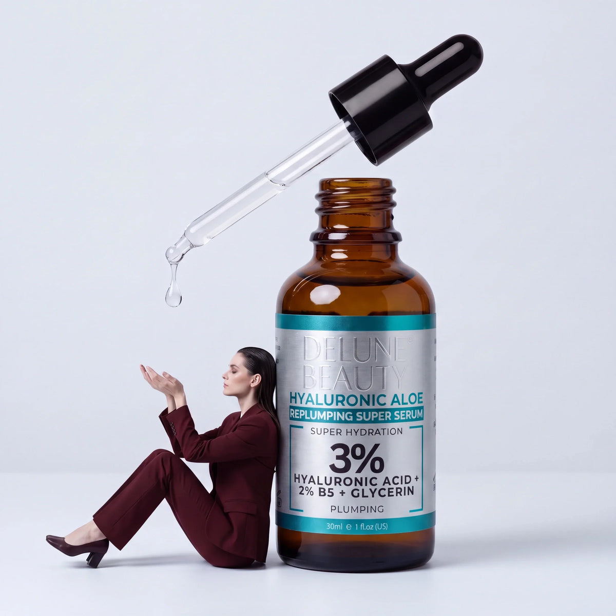 Restaurer : une femme en tailleur bordeaux appuyée contre un flacon géant de sérum Hyaluronic Aloe Delune attrape une goutte qui tombe